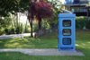 Die blaue Telefonzelle von der Seite fotografiert. Die Zelle steht am Ende eines barrierenfreien Weges inmitten einer Wiese. Im Hintergrund sieht man Bäume und den Weg am Innufer..