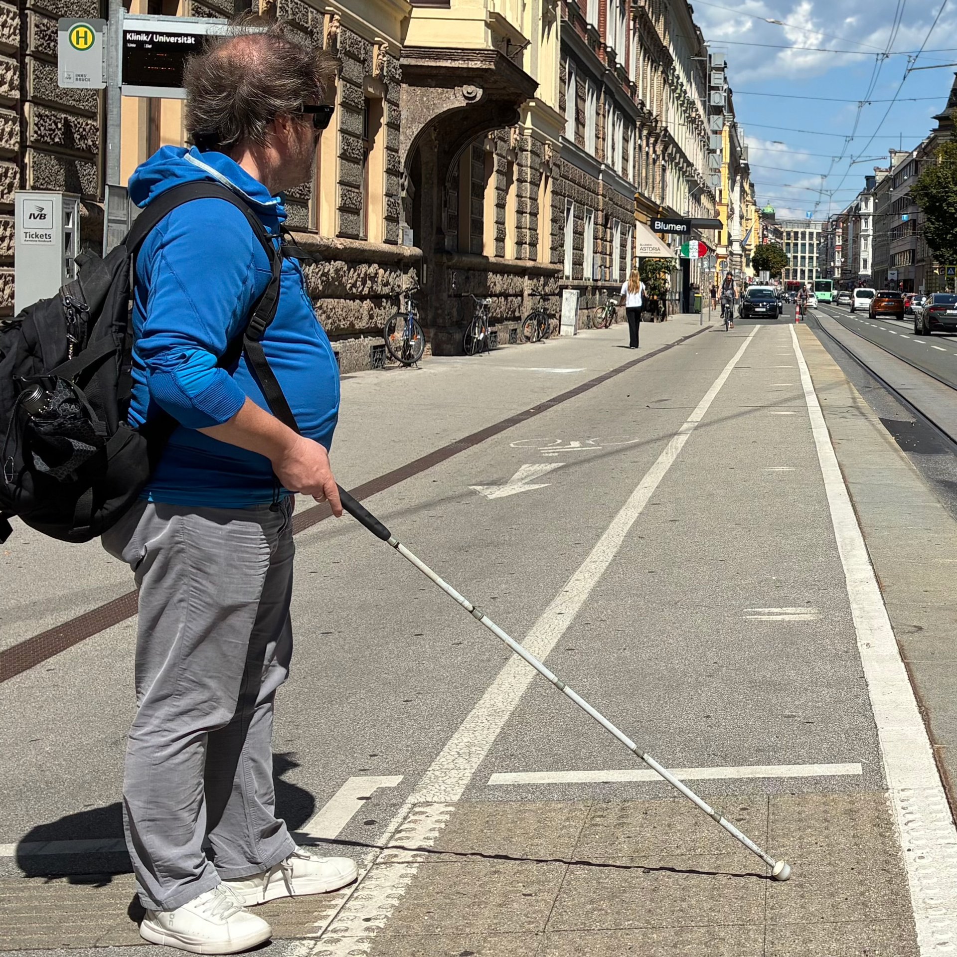 Eine Person steht an einer Straßenbahnhaltestelle und hält einen weißen Langstock auf einer Bodenfläche mit genoppten taktilen Markierungen.