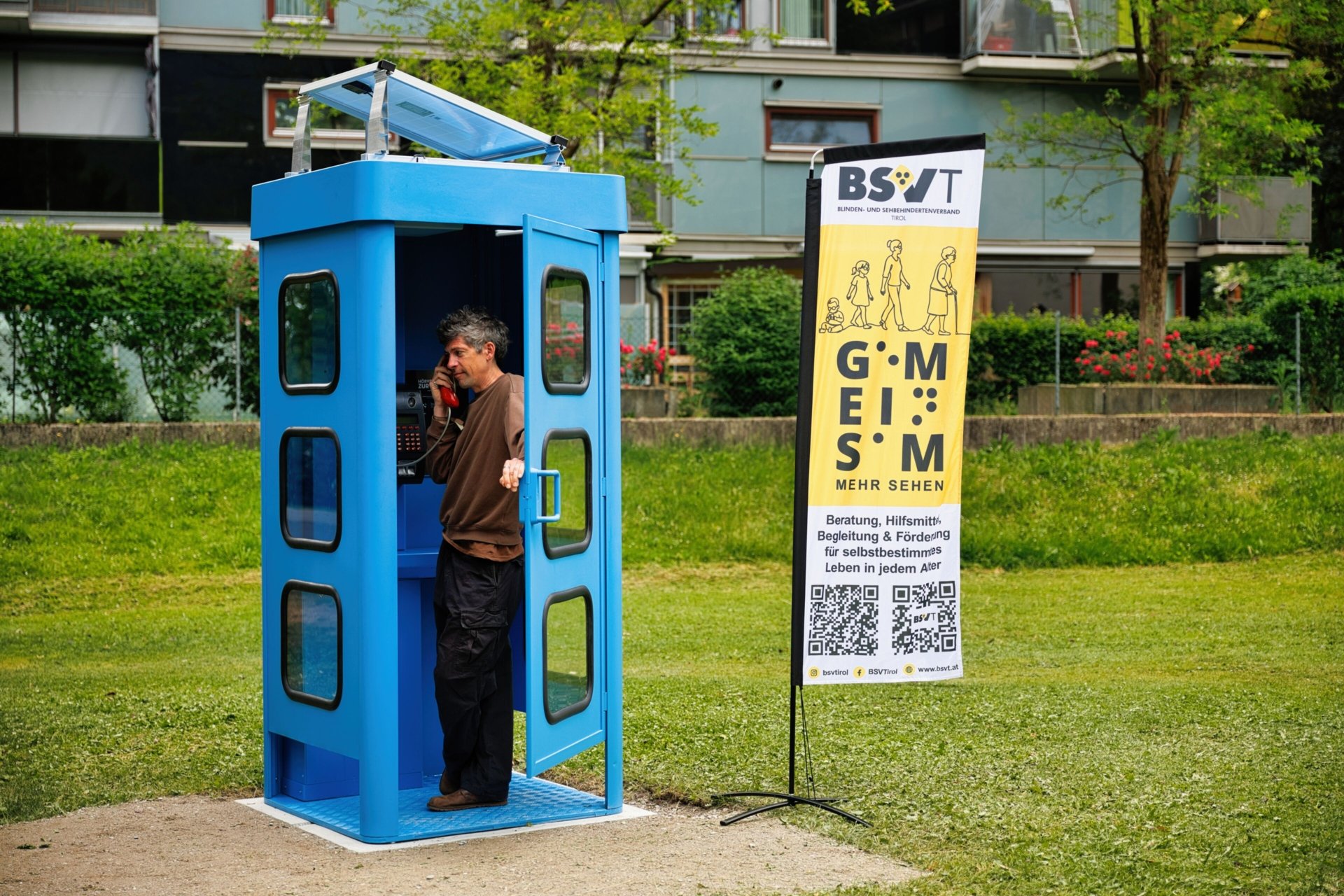 Bosko Gastager steht in der Kunstinstallation „Hörverbindung zur Quelle“, einer umfunktionierten Telefonzelle im öffentlichen Raum, und hört über einen Telefonhörer Klangaufnahmen von Innsbrucker Trinkwasserquellen. Neben der Installation steht ein Informationsbanner des BSVT.