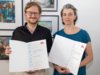Die Kompositions-StipendiatInnen Erwan Borek und Judith Unterpertinger halten ihre Urkunden in der Hand und lächeln in die Kamera. Dahinter sieht man eine Bilderwand. 