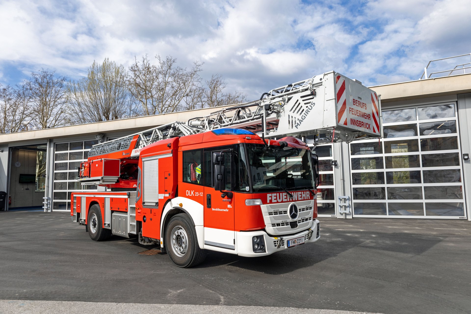 Ein rotes Fahrzeug mit Drehleiter mit Korb steht bei Tageslicht vor dem Gebäude der Berufsfeuerwehr Innsbruck.