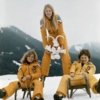 Drei Athletinnen der Olympischen Winterspiele 1976 mit dem Schneemann-Maskottchen "Snowy" auf der Skipiste, zwei davon sitzen auf Rodeln.