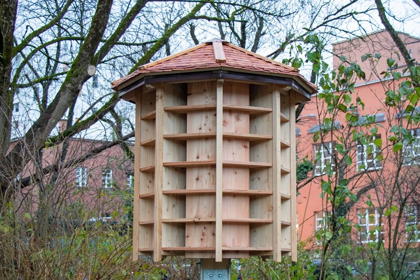 eine achteckige Holzkonstruktion mit vielen einzelnen Fächern und einem Dach, im Hintergrund das Hallenbad Amraser Straße