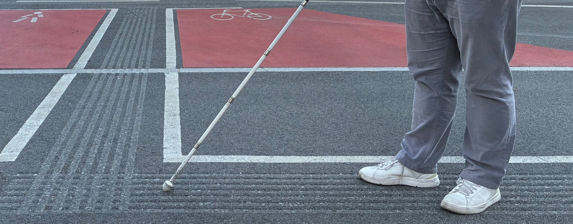 Person mit Langstock orientiert sich an taktilen Bodenleitlinien an einer Straßenquerung. Es sind nur die Beine zu sehen. Der weiße Langstock tastet die gerillte Bodenstruktur ab, die zur sicheren Orientierung für blinde und sehbehinderte Menschen dient.