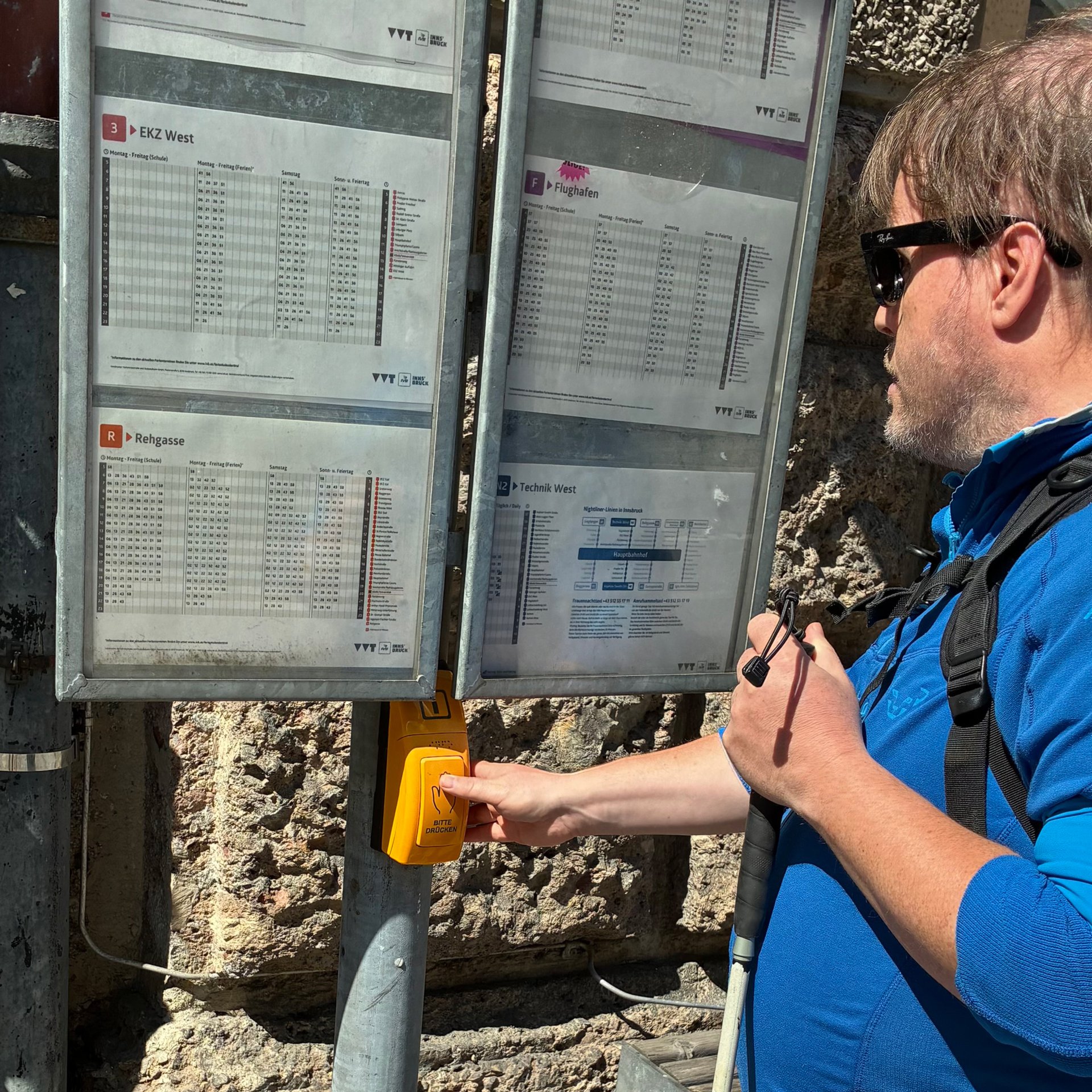 Eine Person steht vor einem Fahrplankasten an einer Haltestelle und drückt einen gelben Informations-Taster an einer Metallstange.