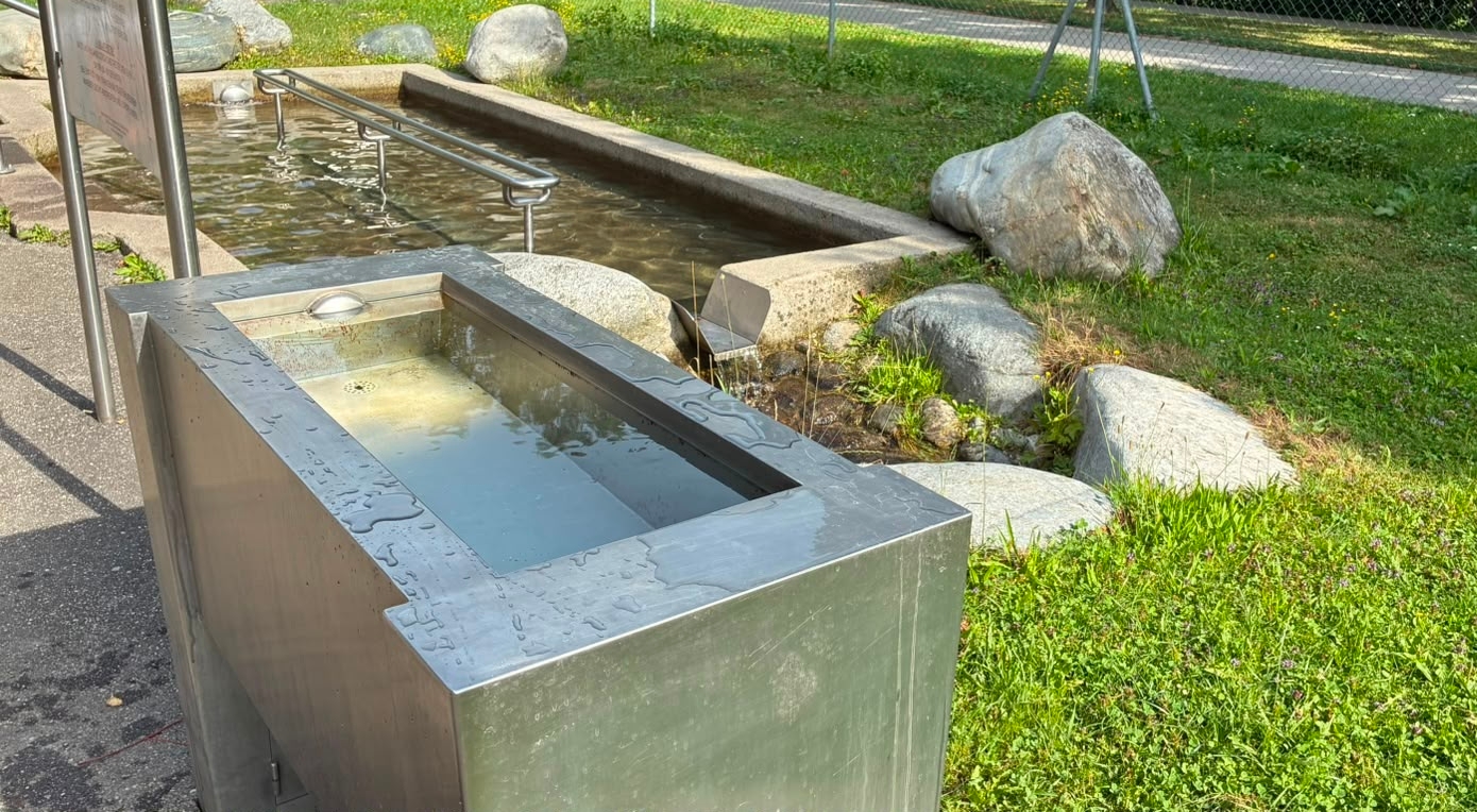 Ein rechteckiges, freistehendes Kneipp-Arm-Badebecken aus Edelstahl, das am Rand eines asphaltierten Weges steht. Der Trog ist mit klarem Wasser gefüllt. Rechts vom Becken befindet sich eine Grünfläche mit großen Flusssteinen. Im Hintergrund ist ein breiteres, flaches Kneipp-Tretbecken mit Edelstahl-Handläufen zu erkennen.