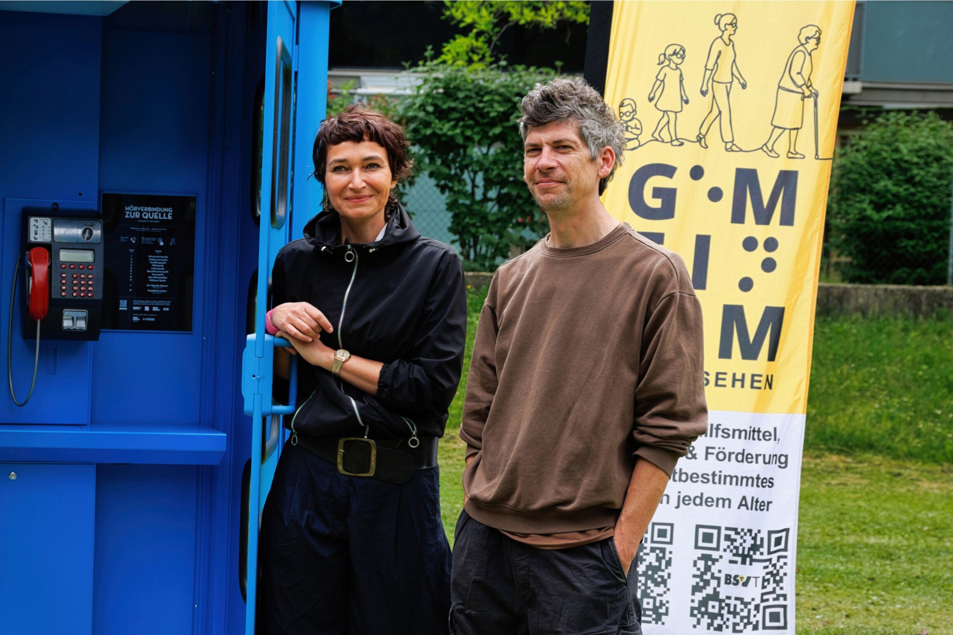 Kata Hinterlechner und Bosko Gastager stehen neben der Kunstinstallation „Hörverbindung zur Quelle“, einer leuchtend blauen, umgestalteten Telefonzelle. Die Tür der Installation ist geöffnet, daneben steht ein Informationsbanner des Blinden- und Sehbehindertenverbands Tirol.