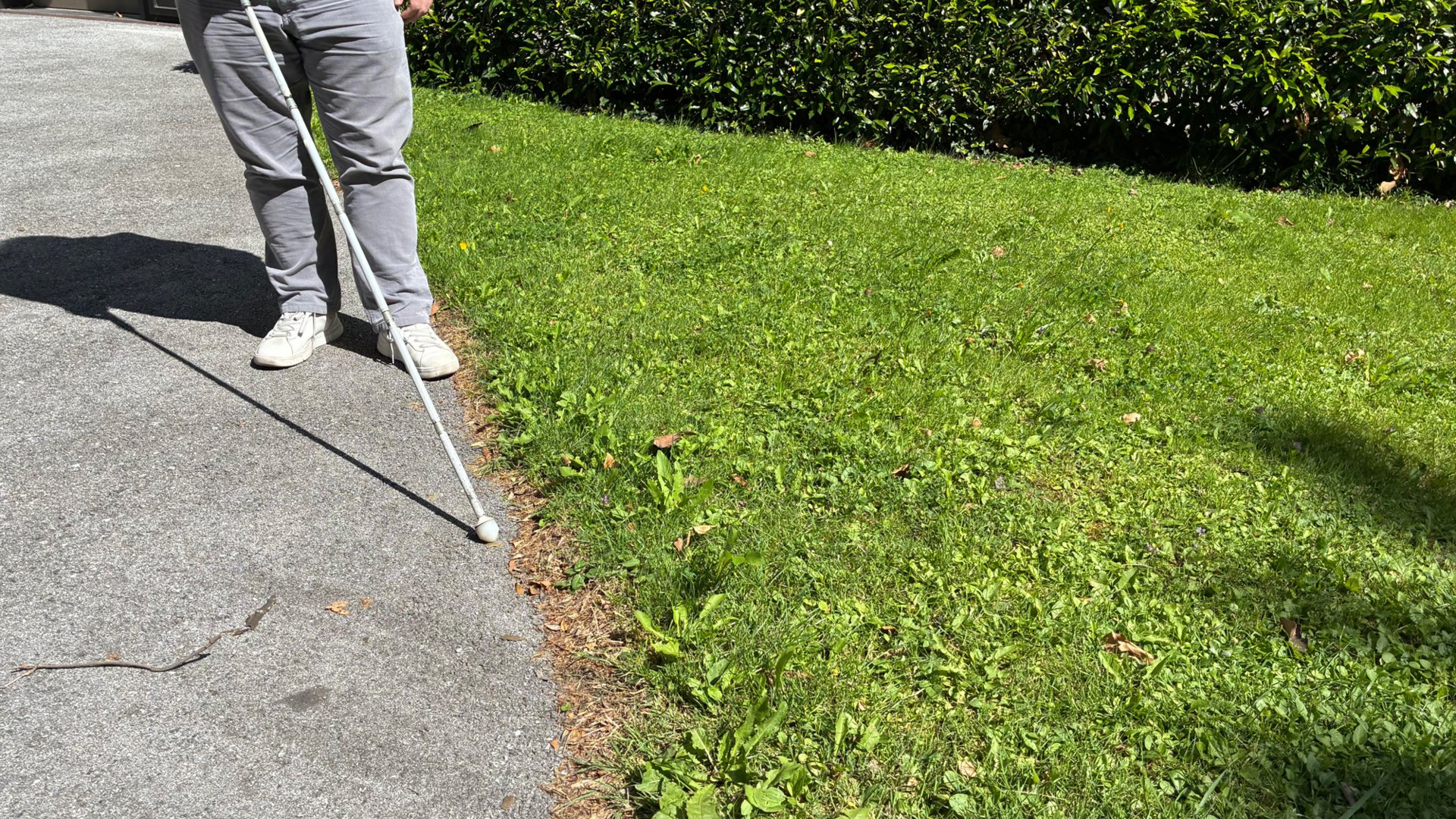 Eine Person tastet mit einem weißen Langstock den Übergang zwischen einem Gehweg und einer angrenzenden Rasenfläche ab.