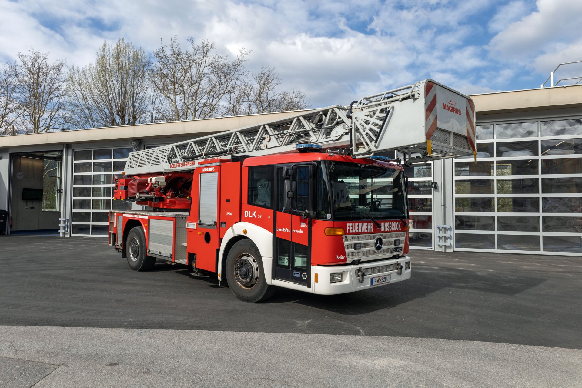 Ein rotes Fahrzeug mit Drehleiter mit Korb steht bei Tageslicht vor dem Gebäude der Berufsfeuerwehr Innsbruck.