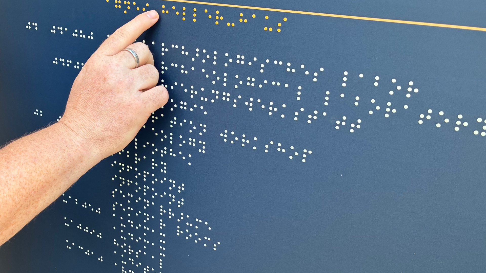 Eine Hand fährt mit dem Zeigefinger über eine dunkelblaue Informationstafel mit Brailleschrift. Die erhabenen Punkte sind in mehreren Reihen angeordnet und können durch Tasten gelesen werden.