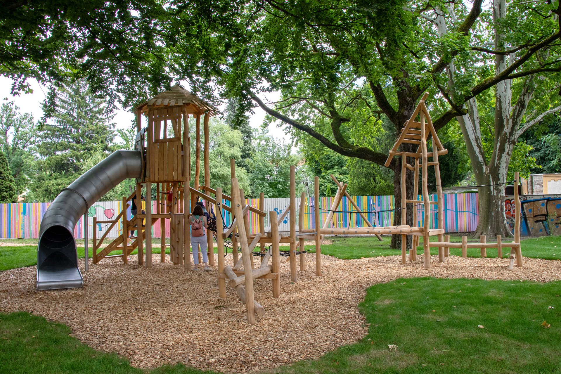 Spielgeräte aus Holz in einem grünen Park, im Hintergrund ein bunt bemalter Zaun